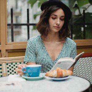 A young woman enjoys reading a book indoors with coffee and a croissant on a relaxing day.